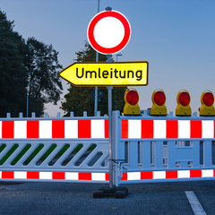 Ein geschlossener Straßenabschnitt mit einer Absperrung und einem Schild, das auf eine Umleitung hinweist. Hinter der Absperrung sind mehrere gelbe Warnleuchten sichtbar. Das Bild vermittelt eine Verkehrswarnung und zeigt eine Umleitung für Fahrzeuge an.