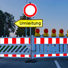 Ein geschlossener Straßenabschnitt mit einer Absperrung und einem Schild, das auf eine Umleitung hinweist. Hinter der Absperrung sind mehrere gelbe Warnleuchten sichtbar. Das Bild vermittelt eine Verkehrswarnung und zeigt eine Umleitung für Fahrzeuge an.