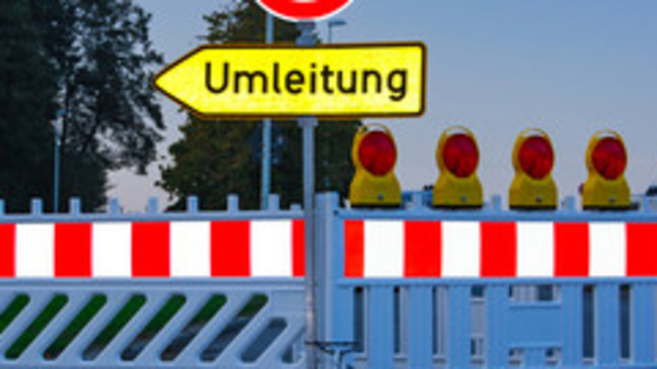 Ein geschlossener Straßenabschnitt mit einer Absperrung und einem Schild, das auf eine Umleitung hinweist. Hinter der Absperrung sind mehrere gelbe Warnleuchten sichtbar. Das Bild vermittelt eine Verkehrswarnung und zeigt eine Umleitung für Fahrzeuge an.