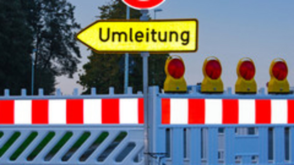 Ein geschlossener Straßenabschnitt mit einer Absperrung und einem Schild, das auf eine Umleitung hinweist. Hinter der Absperrung sind mehrere gelbe Warnleuchten sichtbar. Das Bild vermittelt eine Verkehrswarnung und zeigt eine Umleitung für Fahrzeuge an.