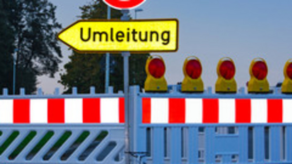 Une barrière de chantier avec un panneau d'interdiction de circulation et un panneau indiquant une déviation. La barrière est rouge et blanche, avec des feux orange allumés au-dessus. L'image montre une route bloquée et informe les automobilistes de prendre un itinéraire alternatif.
