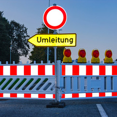 Ein geschlossener Straßenabschnitt mit einer Absperrung und einem Schild, das auf eine Umleitung hinweist. Hinter der Absperrung sind mehrere gelbe Warnleuchten sichtbar. Das Bild vermittelt eine Verkehrswarnung und zeigt eine Umleitung für Fahrzeuge an.