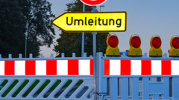 Ein geschlossener Straßenabschnitt mit einer Absperrung und einem Schild, das auf eine Umleitung hinweist. Hinter der Absperrung sind mehrere gelbe Warnleuchten sichtbar. Das Bild vermittelt eine Verkehrswarnung und zeigt eine Umleitung für Fahrzeuge an.
