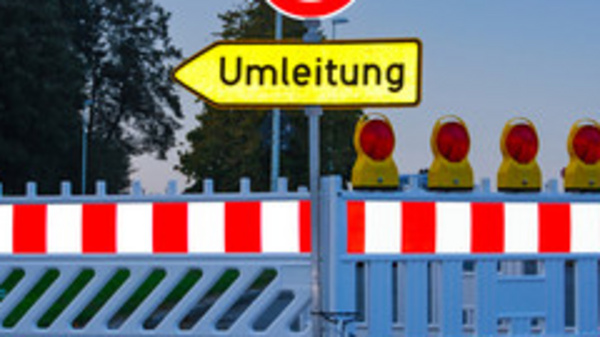 Ein geschlossener Straßenabschnitt mit einer Absperrung und einem Schild, das auf eine Umleitung hinweist. Hinter der Absperrung sind mehrere gelbe Warnleuchten sichtbar. Das Bild vermittelt eine Verkehrswarnung und zeigt eine Umleitung für Fahrzeuge an.