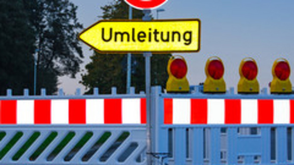 A roadblock is featured with a red and white barrier, and a circular road sign indicating 'no entry.' There is a yellow sign pointing to the right labeled "Umleitung," which means "detour" in German. Several warning lights are positioned at the top of the barrier.