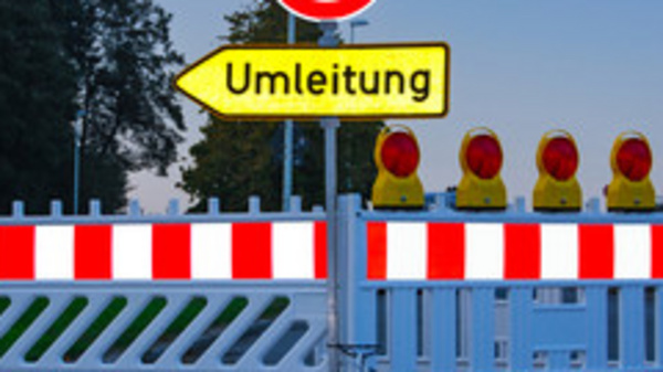 A roadblock is featured with a red and white barrier, and a circular road sign indicating 'no entry.' There is a yellow sign pointing to the right labeled "Umleitung," which means "detour" in German. Several warning lights are positioned at the top of the barrier.