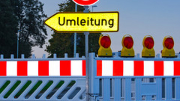 A roadblock is featured with a red and white barrier, and a circular road sign indicating 'no entry.' There is a yellow sign pointing to the right labeled "Umleitung," which means "detour" in German. Several warning lights are positioned at the top of the barrier.