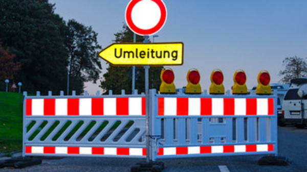 A roadblock is featured with a red and white barrier, and a circular road sign indicating 'no entry.' There is a yellow sign pointing to the right labeled "Umleitung," which means "detour" in German. Several warning lights are positioned at the top of the barrier.