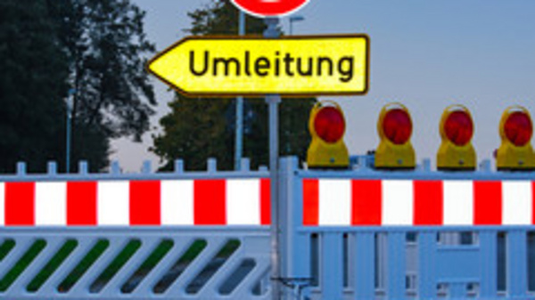 Ein geschlossener Straßenabschnitt mit einer Absperrung und einem Schild, das auf eine Umleitung hinweist. Hinter der Absperrung sind mehrere gelbe Warnleuchten sichtbar. Das Bild vermittelt eine Verkehrswarnung und zeigt eine Umleitung für Fahrzeuge an.