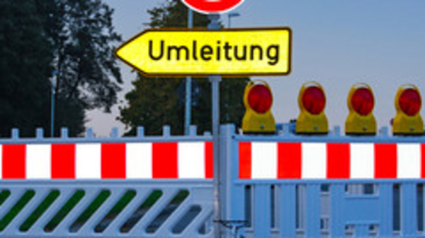 Ein geschlossener Straßenabschnitt mit einer Absperrung und einem Schild, das auf eine Umleitung hinweist. Hinter der Absperrung sind mehrere gelbe Warnleuchten sichtbar. Das Bild vermittelt eine Verkehrswarnung und zeigt eine Umleitung für Fahrzeuge an.