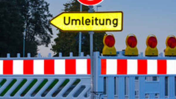 Ein geschlossener Straßenabschnitt mit einer Absperrung und einem Schild, das auf eine Umleitung hinweist. Hinter der Absperrung sind mehrere gelbe Warnleuchten sichtbar. Das Bild vermittelt eine Verkehrswarnung und zeigt eine Umleitung für Fahrzeuge an.