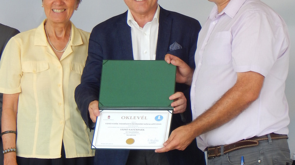 Three individuals stand together, smiling. The center figure holds a certificate in one hand and is shaking hands with another person. The first individual on the left wears a light yellow shirt, while the third person on the right is dressed in a light purple shirt.