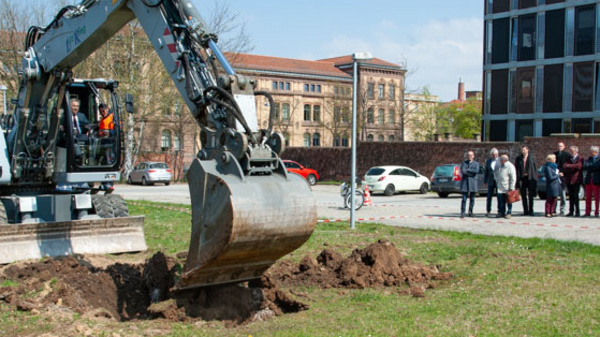 An excavator digs into the ground on a grassy area, with a crowd of people watching in the background. In the distance, a modern building stands alongside older architectural structures. The setting appears to be an outdoor event or ceremony.