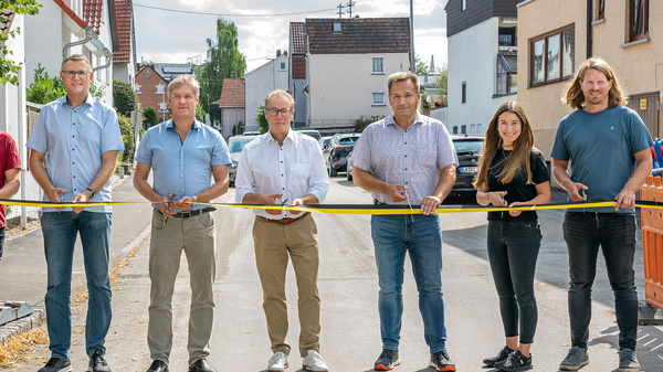 Eine Gruppe von sieben Personen steht mit einer Absperrband auf einer Straße. Die Personen sind in formeller Kleidung. Im Hintergrund sind Wohnhäuser und ein Baustellenbereich sichtbar. Die Szene vermittelt eine Botschaft der Straßensperrung oder einer offiziellen Eröffnung.