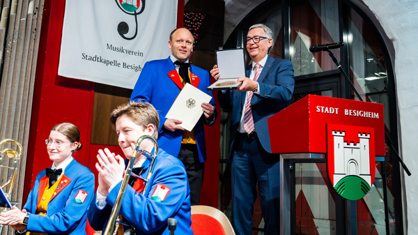 Bei einer Zeremonie in Besigheim übergibt ein Mann Anerkennung an einen Musiker in Uniform. Im Hintergrund sitzen weitere Musiker, darunter eine Frau mit einem Instrument. Eine Flagge der Stadt sowie ein Podium mit dem städtischen Logo sind sichtbar.