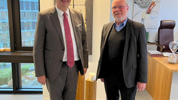 Zwei Männer stehen in einem modernen Büroraum. Der eine trägt einen Anzug mit einer Krawatte, der andere einen dunklen Anzug und Pulli. Im Hintergrund ist ein Schreibtisch und ein Fenster mit Blick nach draußen zu sehen.