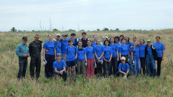 Eine Gruppe von etwa 30 Menschen steht lächelnd auf einer offenen Wiese. Die meisten tragen blaue T-Shirts. Die Umgebung ist ländlich, mit etwas hohem Gras und wenigen Bäumen im Hintergrund. Der Himmel ist bewölkt.