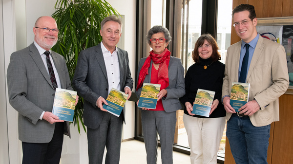 A group of five professionals stands together in a well-lit office, each holding a book. They are engaged in conversation, showcasing their publications related to a specific topic. The atmosphere is collaborative and emphasizes the importance of the books they are presenting.