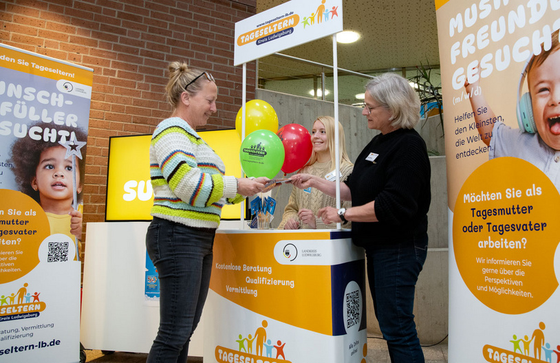 Eine Frau übergibt bunte Ballons an eine andere Frau an einem Stand. Im Hintergrund stehen weitere Personen. Der Stand informiert über die Möglichkeiten, als Tagesmutter zu arbeiten. Plakate mit Aufschriften bewerben die Suche nach Tageseltern und Informationen zur Qualifizierung.