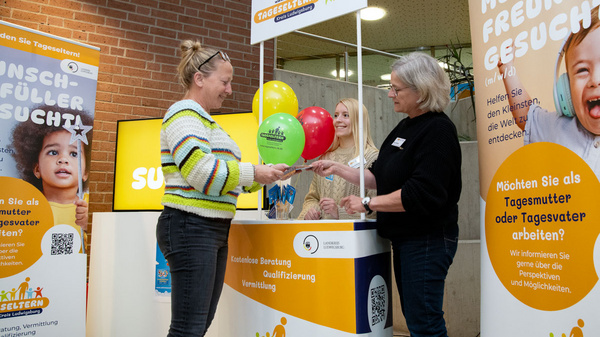 Eine Frau übergibt bunte Ballons an eine andere Frau an einem Stand. Im Hintergrund stehen weitere Personen. Der Stand informiert über die Möglichkeiten, als Tagesmutter zu arbeiten. Plakate mit Aufschriften bewerben die Suche nach Tageseltern und Informationen zur Qualifizierung.