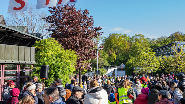 Mehrere Menschen versammeln sich an einem sonnigen Tag unter einem großen "START"-Schild. Die Umgebung ist grün, mit Bäumen und Gebäuden im Hintergrund. Die Teilnehmer wirken bereit, an einer Veranstaltung oder einem Wettlauf teilzunehmen.