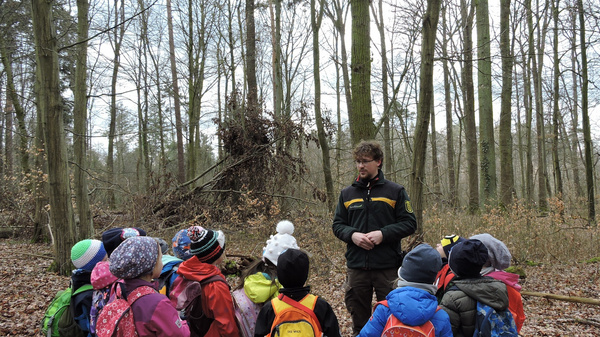 Un educatore spiega ai bambini in un bosco, mentre stanno attorno a lui. I bambini indossano giacche colorate e zaini, osservando attentamente. Lo sfondo mostra alberi spogli, tipici di una giornata invernale.