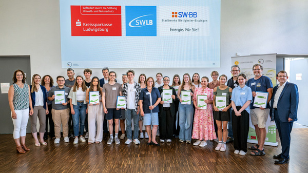 Die Gruppe von jungen Personen steht zusammen in einem Raum, jeder hält eine Urkunde in der Hand. Im Hintergrund sind Logos von unterstützenden Institutionen zu sehen. Die Stimmung ist feierlich und die Teilnehmer wirken stolz auf ihre Leistungen.