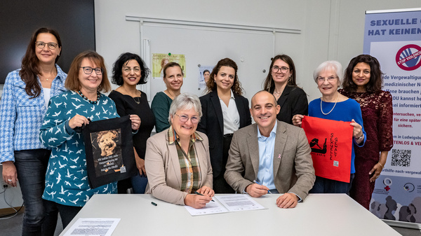 Eine Gruppe von Frauen und einem Mann steht um einen Tisch, auf dem zwei Personen Verträge unterzeichnen. Im Hintergrund ist ein Plakat zu sehen, das gegen sexuelle Gewalt richtet. Alle Beteiligten zeigen Engagement für das Thema und halten Flaggen oder Tücher in der Hand.