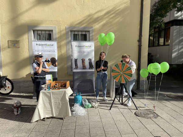 Due persone sono presenti in un evento all'aperto, una delle quali tiene dei palloncini verdi. Un'altra è accanto a una ruota della fortuna. In primo piano, un tavolo è coperto con vari materiali promozionali e una piccola cagnolino è seduto vicino.