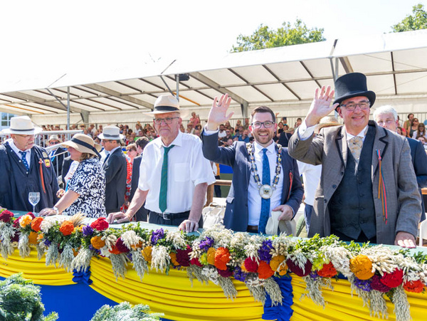 24.08.2024 - Beim Schäferlauf mit Bürgermeister Jens Hübner (mitte) & Ministerpräsident Winfried Kretschmann (li.) Foto: Andreas Essig Gruppenszene bei einem Festakt im Freien. Mehrere Männer in Anzügen und Hüten stehen auf einer Bühne, einige winken dem Publikum zu. Im Hintergrund sind Menschenmengen und bunte Dekorationen zu sehen.