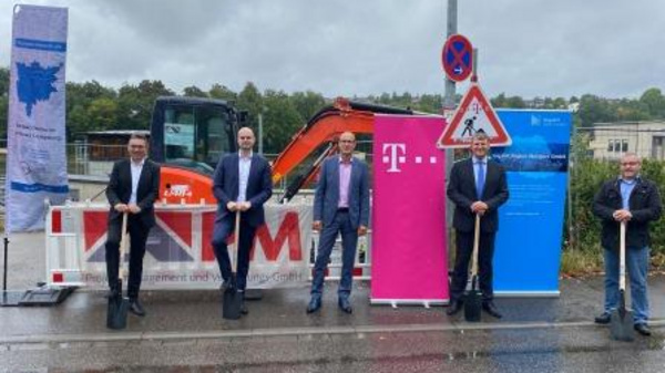 A group of five individuals stands in front of a large orange excavator, holding shovels. They are participating in a ground-breaking ceremony, with banners and a construction sign visible in the background. The atmosphere suggests a celebration of a new development project.