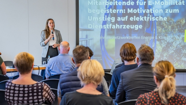 Eine Frau steht vor einem Publikum und spricht über E-Mobilität. Im Hintergrund ist eine Präsentation mit dem Titel „Mitarbeitende für E-Mobilität begeistern: Motivation zum Umstieg auf elektrische Dienstfahrzeuge“ zu sehen. Das Publikum sitzt aufmerksam und hört zu. (Text KI generiert)