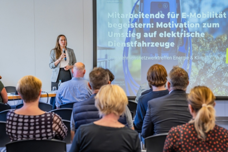 Eine Frau steht vor einem Publikum und spricht über E-Mobilität. Im Hintergrund ist eine Präsentation mit dem Titel „Mitarbeitende für E-Mobilität begeistern: Motivation zum Umstieg auf elektrische Dienstfahrzeuge“ zu sehen. Das Publikum sitzt aufmerksam und hört zu. (Text KI generiert)