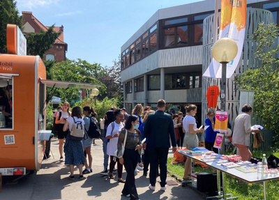 Un gruppo di persone si riunisce all'aperto vicino a un food truck arancione, mentre altri visitano una tavola informativa con materiali. Si possono vedere edifici moderni sullo sfondo e una giornata di sole. L'atmosfera è vivace e sociale.