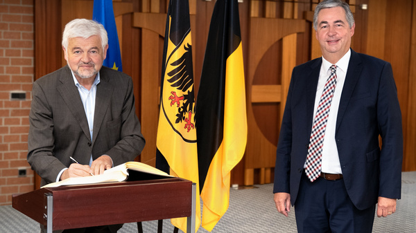 An official event featuring two men in business attire. One man is signing a book at a table, while the other stands beside him, smiling. Behind them are two flags, one with the European Union emblem and the other with the German eagle. The setting appears formal.