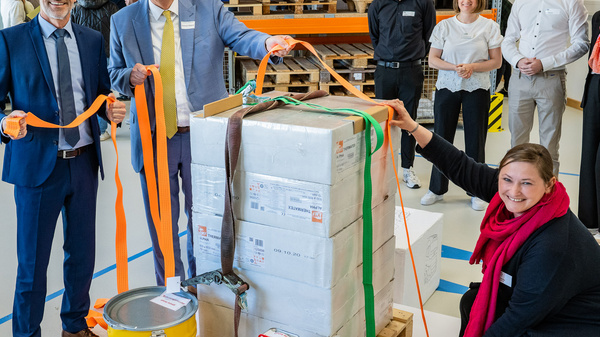 Zwei Männer in Anzügen, einer mit Brille, stehen neben einem großen, verpackten Paket auf einer Palette. Sie halten farbige Bänder, während eine Frau mit rotem Schal auf dem Boden sitzt und lächelt. Im Hintergrund stehen mehrere Personen, die die Szene beobachten.