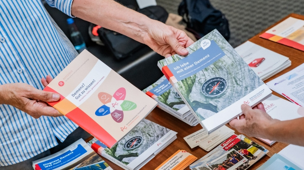 Zwei Personen halten jeweils ein Buch in den Händen, während sie an einem Tisch mit verschiedenen Broschüren und Flyern stehen. Die Bücher sind thematisch gestaltet und scheinen Informationsmaterial zu sein. Der Tisch ist ordentlich mit mehreren Druckerzeugnissen bedeckt. (Text KI generiert)