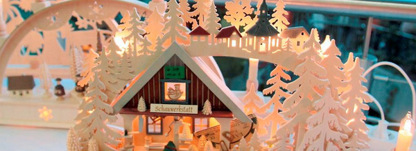 Eine filigrane Holzkrippe mit einem kleinen Häuschen, umgeben von vielen Baumwipfeln. Das Dach des Hauses ist mit Schneeflocken dekoriert. Beleuchtung erzeugt eine gemütliche Atmosphäre. Vor dem Haus stehen Figuren, die eine winterliche Szene darstellen.