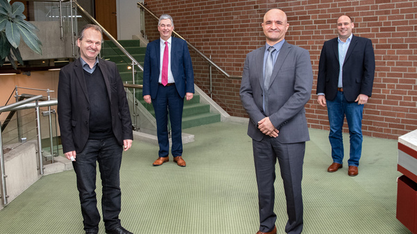 Vier Männer stehen in einem Innenraum mit Pflanzen und einer Treppe im Hintergrund. Sie lächeln und sind formell gekleidet. Im Vordergrund sind sie gleichmäßig verteilt, jeder hat eine entspannte Haltung. Die Atmosphäre wirkt freundlich und professionell. (Text KI generiert)