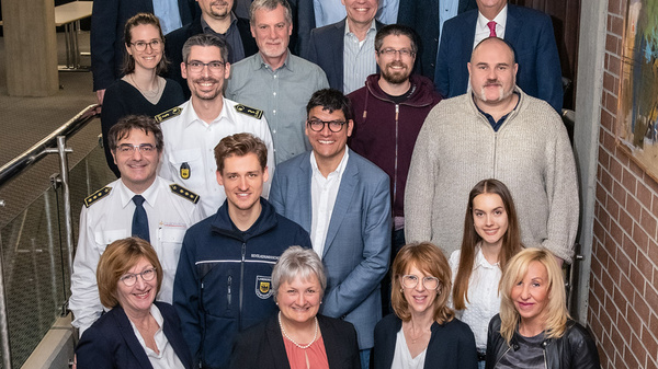 Eine Gruppe von 18 Personen steht auf einer Treppe und posiert für ein Foto. Sie sind in formeller und geschäftlicher Kleidung gekleidet, und es gibt eine Mischung aus Männern und Frauen unterschiedlichen Alters. Im Hintergrund sind moderne architektonische Elemente zu sehen.