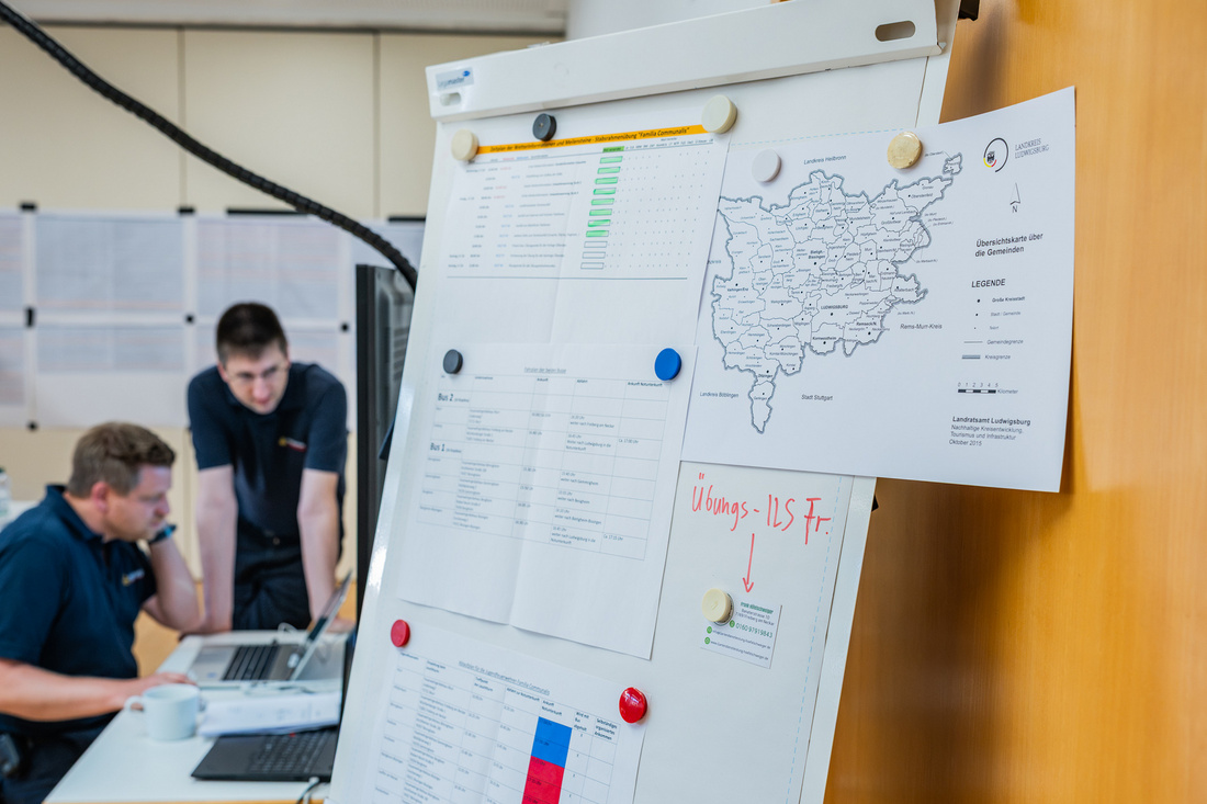 Ein Flipchart in einem Büroraum, auf dem mehrere Arbeitsblätter und eine Karte befestigt sind. Zwei Personen arbeiten am Tisch im Hintergrund an Laptops, konzentriert auf ihre Aufgaben. Die Atmosphäre wirkt geschäftig und kollaborativ.