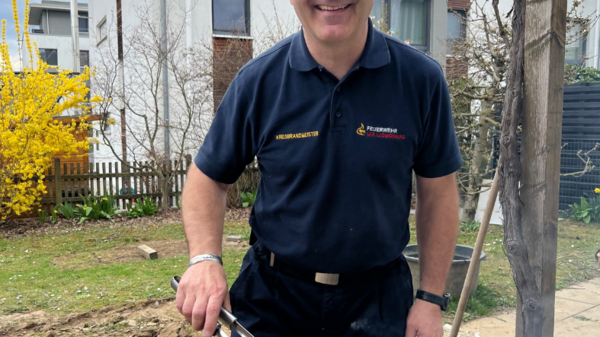 Ein Mann mit Brille steht lächelnd neben einem Grill, auf dem Würstchen und eine Folienverpackung liegen. Er hält eine Grillzange in der Hand, bei sonnigem Wetter im Garten. Im Hintergrund sind Bäume und ein helles Gebäude zu sehen.