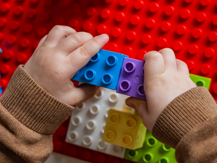 Ein Kind baut mit bunten Lego-Steinen. Es hält zwei Steine in den Händen: einen blauen und einen lila, die gerade zusammengefügt werden. Die Oberfläche, auf der gespielt wird, besteht aus Lego-Platten in verschiedenen Farben.