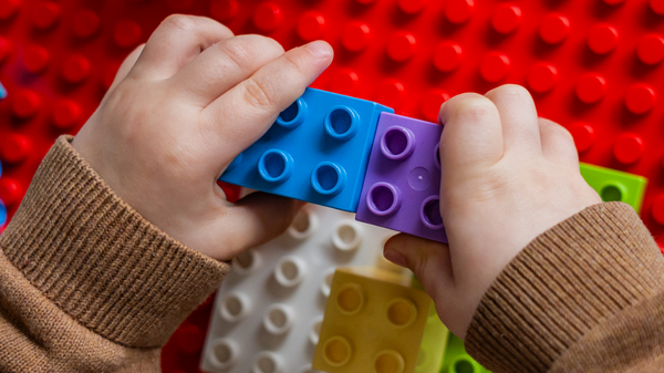 Ein Kind baut mit bunten Lego-Steinen. Es hält zwei Steine in den Händen: einen blauen und einen lila, die gerade zusammengefügt werden. Die Oberfläche, auf der gespielt wird, besteht aus Lego-Platten in verschiedenen Farben.