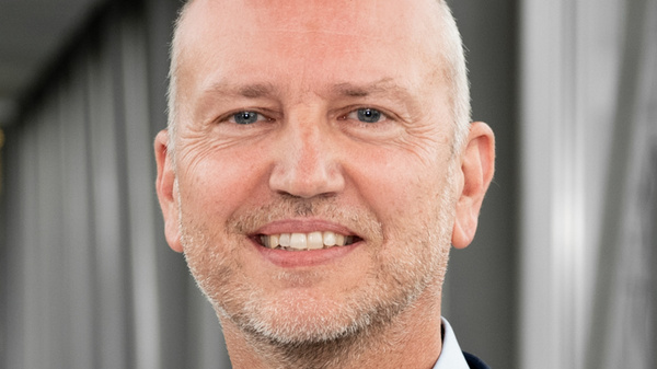 Ein lächelnder Mann mit kurzen, grauen Haaren trägt einen blauen Anzug und ein hellblaues Hemd. Er steht vor einer modernen Architektur, die unscharf im Hintergrund zu sehen ist. Der Ausdruck vermittelt Freundlichkeit und Professionalität.