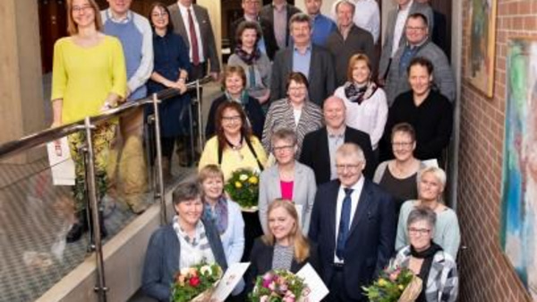 A diverse group of approximately 30 people is gathered in a well-lit indoor space, some holding bouquets of flowers. They are smiling and appear to be celebrating a special occasion. The environment features modern architecture and decorative artwork.