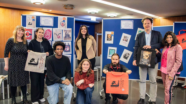 Eine Gruppe von sechs Personen steht in einem Raum, einige halten Stoffbeutel. Im Hintergrund sind Plakate und Präsentationen an Wänden zu sehen. Die Atmosphäre wirkt freundlich und kreativ, was auf eine Veranstaltung oder Ausstellung hinweist.