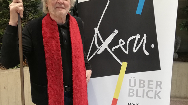 A man stands next to an exhibition banner featuring the name "Wolfgang Kern" and the title "Überblick." He wears a red scarf and holds a pole. The banner details the exhibition dates from February 25 to March 19, 2021, at Kunst im Kreishaus.