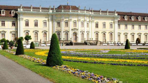 A large, elegant palace with a light yellow facade stands at the center, surrounded by manicured gardens. Flower beds with colorful blooms and neatly trimmed hedges line a pathway leading to the building. Benches for seating are visible on the side.