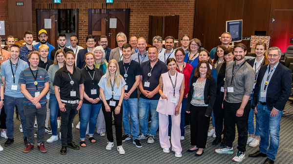 Eine große Gruppe von etwa 50 Personen steht lächelnd zusammen in einem Konferenzraum. Die Teilnehmer tragen unterschiedliche Kleidung und Namensschilder. Im Hintergrund sind Wände mit Kunstwerken und Veranstaltungen sichtbar, die auf eine professionelle Zusammenkunft hinweisen.
